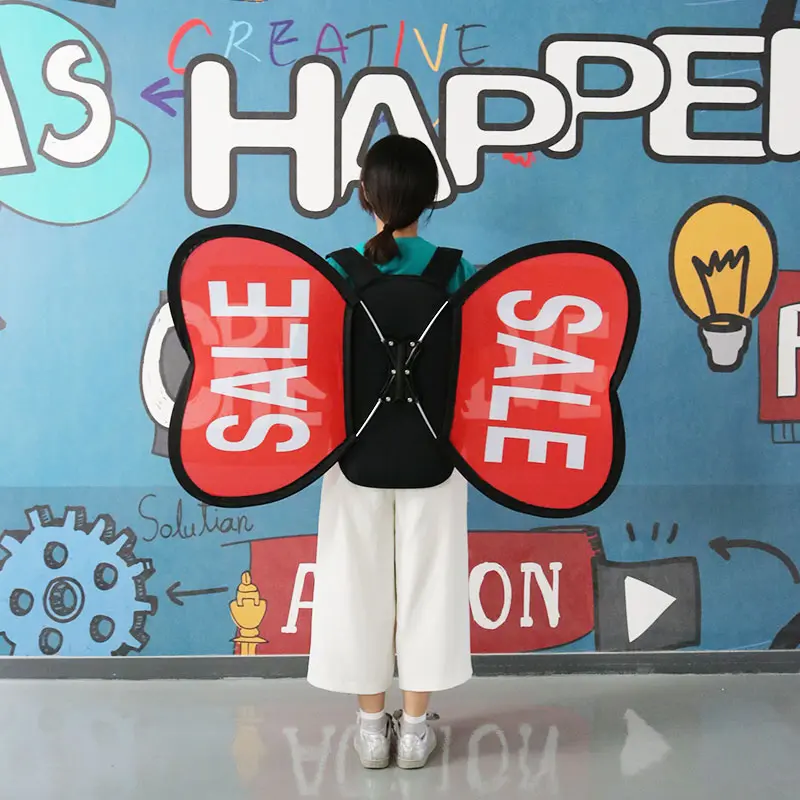 Creative marketing application of custom promotional backpack with red butterfly-shaped banners featuring bold 'SALE' text. Urban-style presentation against graffiti wall background with 'CREATIVE HAPPEN' artwork, highlighting mobile advertising versatility for retail environments.