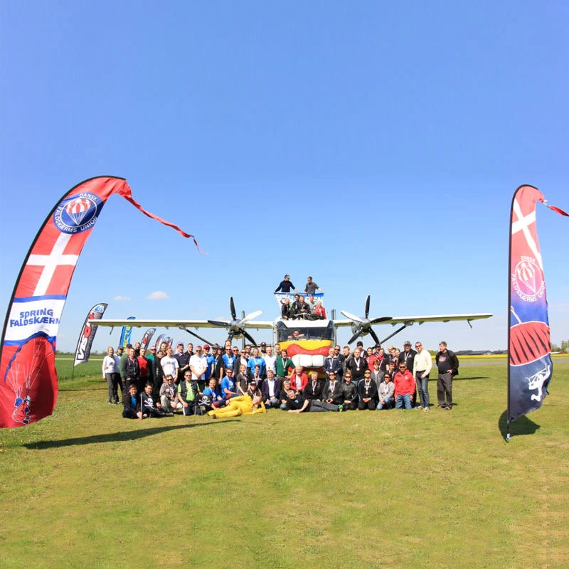 Group photo in front of a twin-propeller airplane on a grassy field, flanked by two vertical bali banner flags
