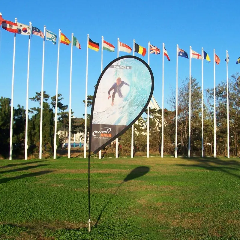 R Banner outdoor event setup: Teardrop-shaped banner with surfer graphic on green lawn. Multiple national flags in background demonstrate international exhibition use with carbon composite pole stability.