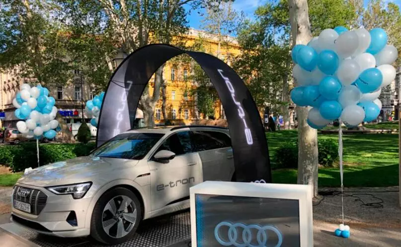 Silver Audi e-tron at outdoor auto showcase with black arch stand banner display, blue/white balloon columns, and Audi-branded signage in a park setting.