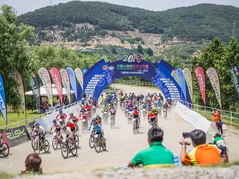 2018 China Mountain Bike Open start line: blue archway, sponsor feathe flags, and cyclists in action with dust trails.