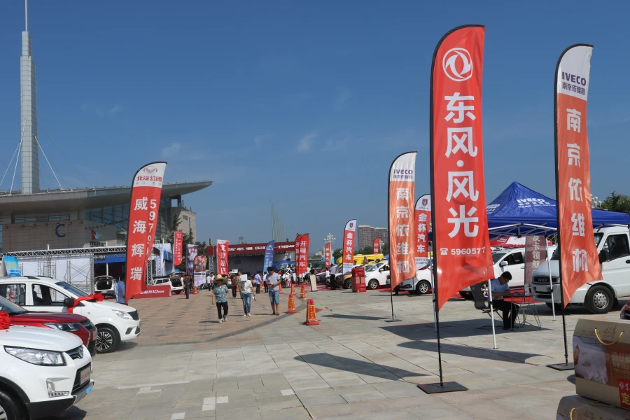 Red automotive blade flags at sunny car expo with crowds, vehicles, and blue sky