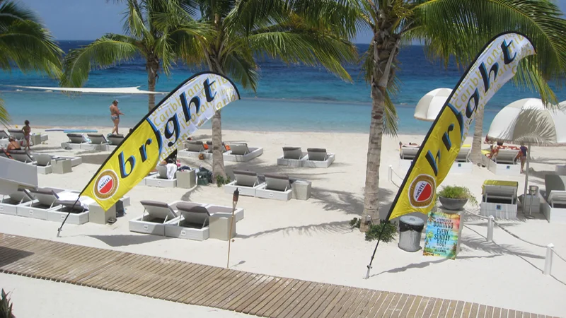 Tall yellow swooper flags on beach with lounge chairs and palm trees, ocean in background.