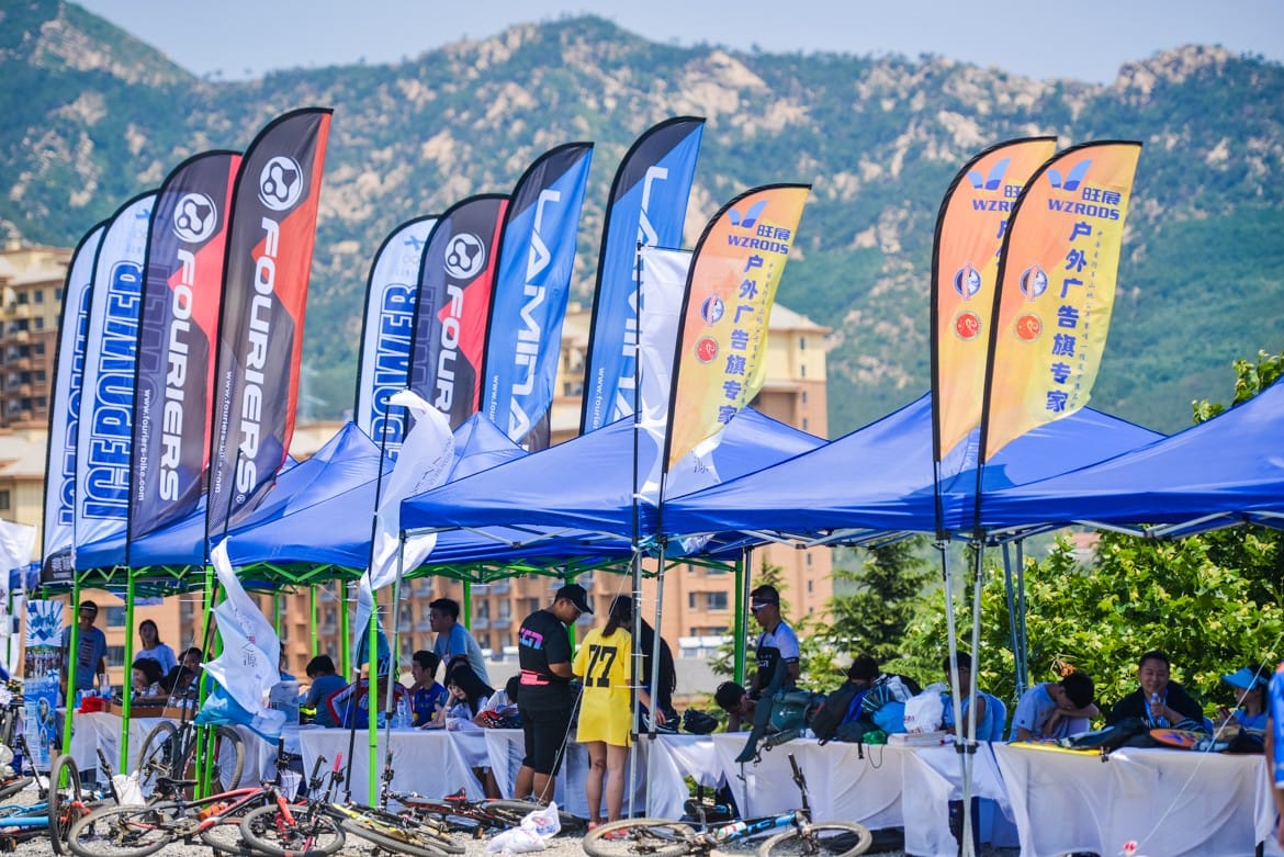 Outdoor event with WZROOS advertising feather flags, tents, event signage against mountain scenery.