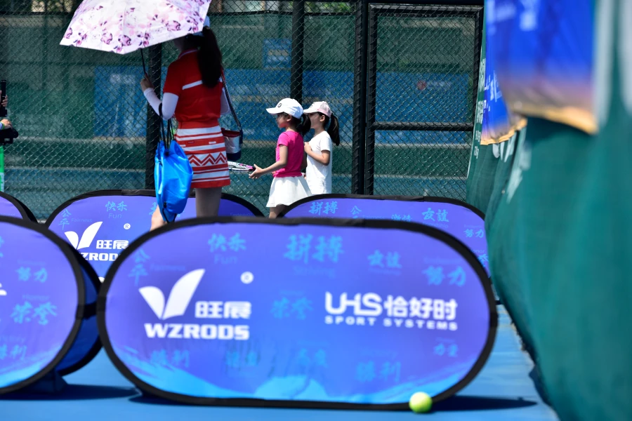 Outdoor tennis event scene with blue sideline banners (WZRODS, UHS), people (including kids) in background, sunny day.