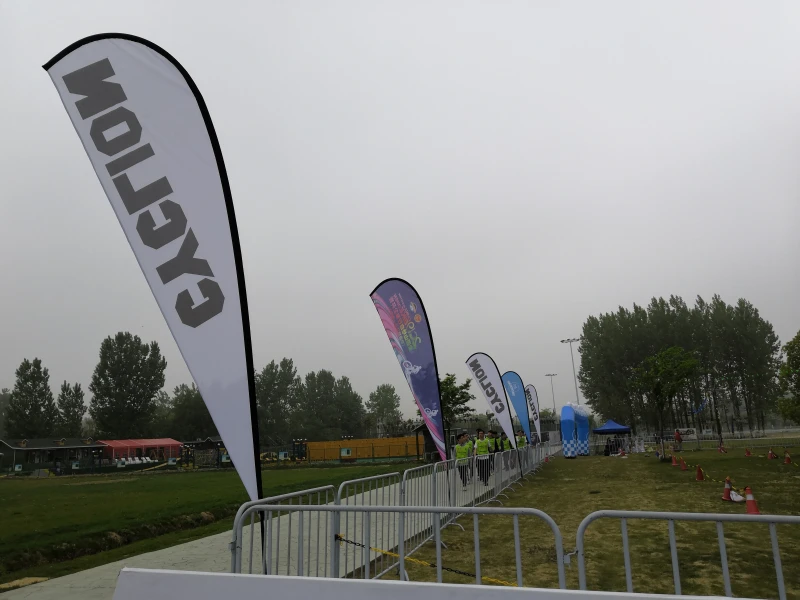 Black 'CYClION' teardrop flag with white border among colorful event teardrop banners near metal railing