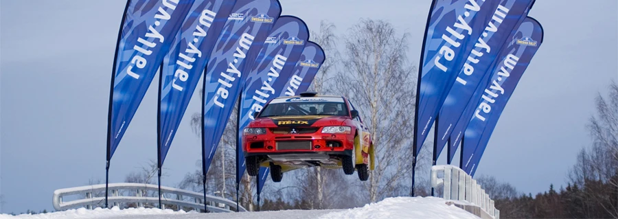 Red rally car jumping over snow with teardrop flags marked 'rally.vm' lining the snowy track