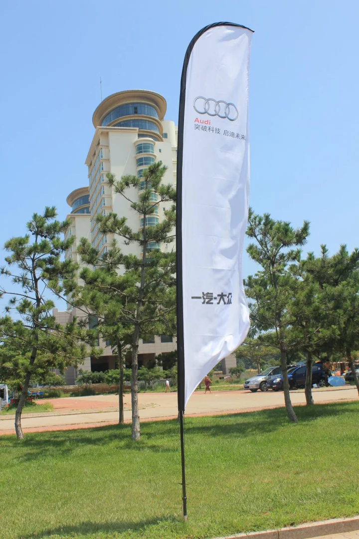 沙滩旗，展示奥迪标志及标语，背景为草地，树木，现代建筑